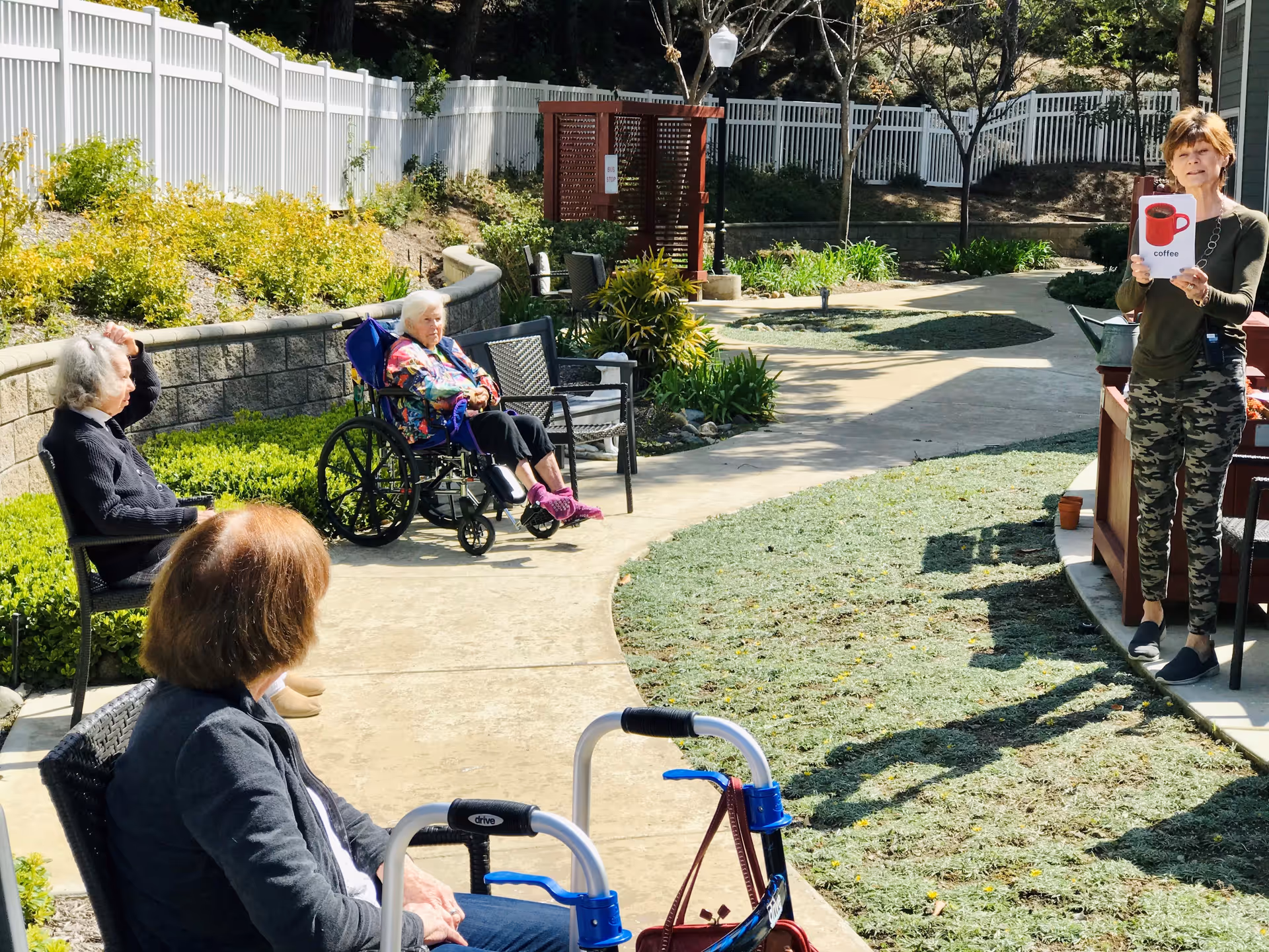 An outdoor garden area at a senior living facility with three elderly women seated along a curved concrete pathway. One woman is in a wheelchair, another is seated on a chair, and a third is partially visible in the foreground. A woman stands on the right side holding a sign with a red coffee cup and the word 'coffee'. The area is surrounded by greenery, a white fence, and trees.