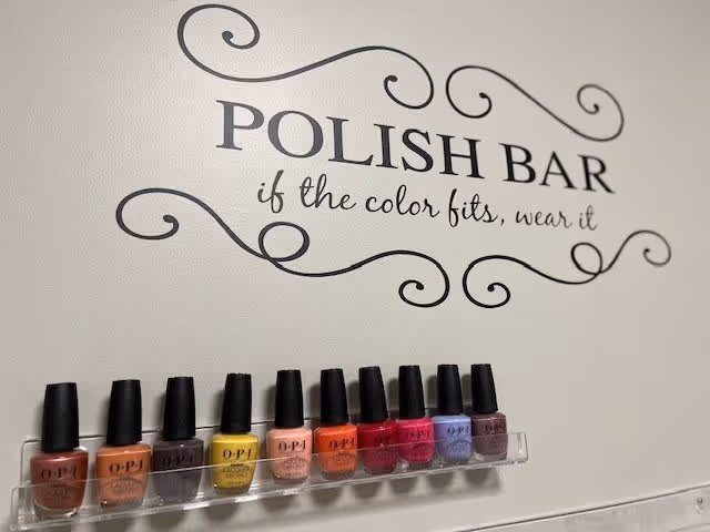 A wall with decorative text that reads 'POLISH BAR if the color fits, wear it' above a clear shelf holding ten bottles of nail polish in various colors.