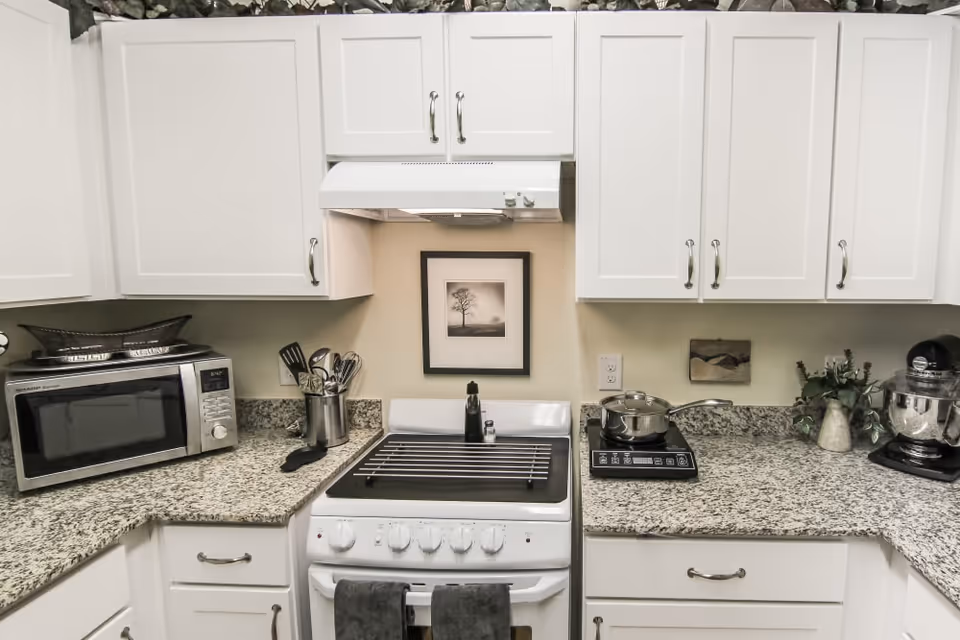 A kitchen with white cabinets and granite countertops. There is a microwave on the left countertop, a stove with a rack on top in the center, and a pot on an electric burner on the right countertop. Kitchen utensils are stored in a container next to the stove, and a stand mixer and a small plant are on the right side. A framed black and white picture of a tree hangs on the wall above the stove.