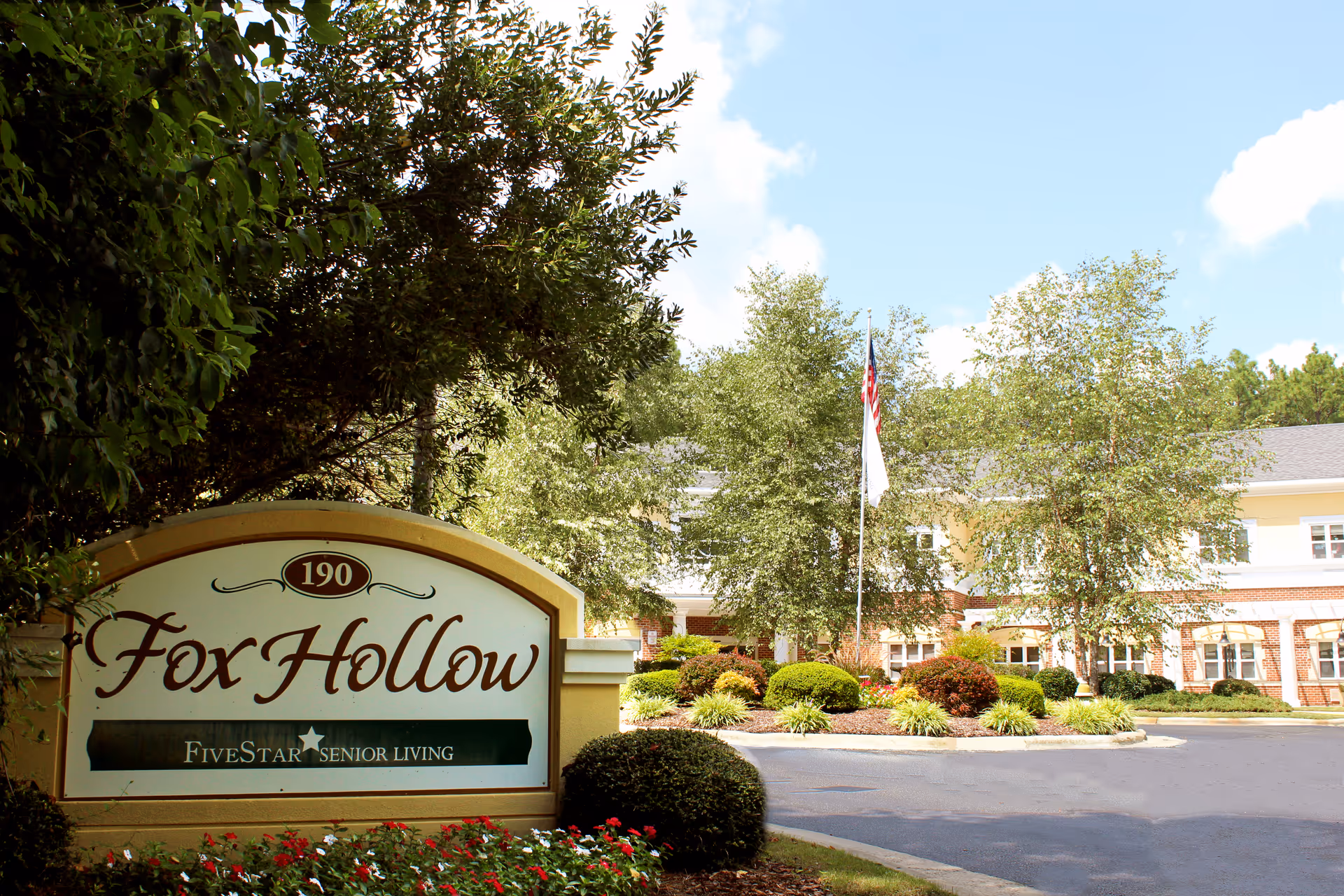 Entrance sign for Fox Hollow Senior Living Community with landscaping and a building in the background under a partly cloudy sky.