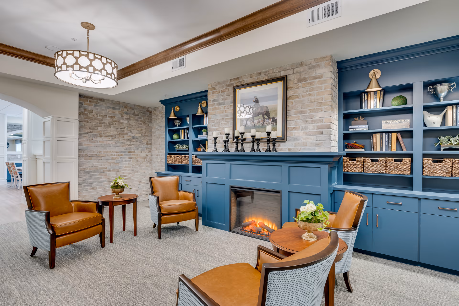 A cozy sitting area with four brown leather chairs arranged around two small wooden tables with flower arrangements. The background features a blue built-in bookshelf and a fireplace with a brick surround. Above the fireplace is a framed picture of a horse. The room has a modern light fixture hanging from the ceiling and a carpeted floor.