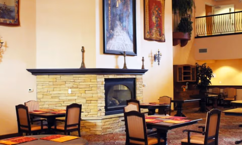 Interior view of a senior living facility dining area with multiple tables and chairs arranged around a stone fireplace. The walls are decorated with framed artwork and sconces, and there is a balcony with a railing visible on the upper level.