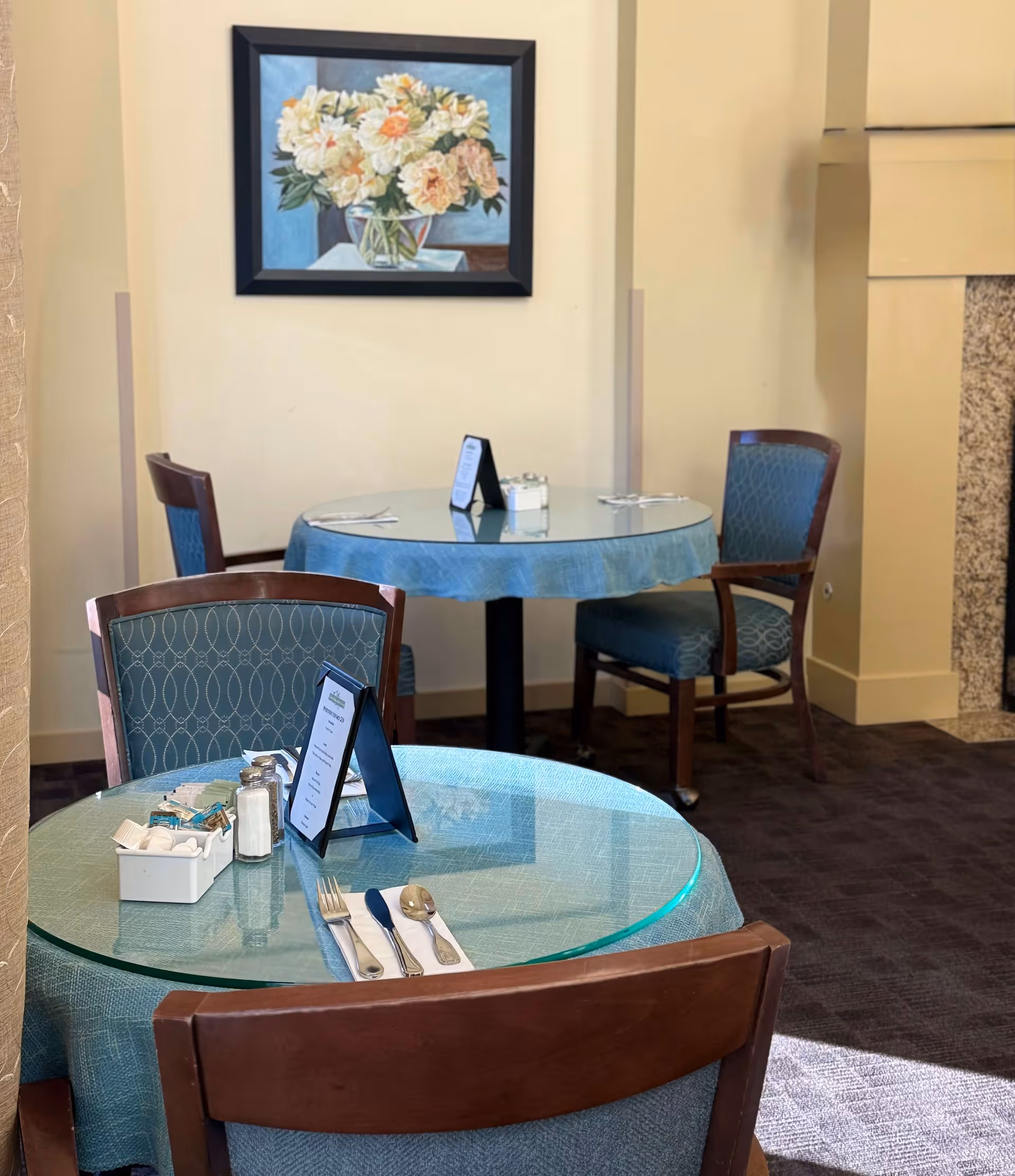 Two round dining tables with glass tops and blue tablecloths, each set with silverware, napkins, salt and pepper shakers, and small menu stands. The tables are surrounded by wooden chairs with blue patterned upholstery. A framed painting of a vase with white and peach flowers hangs on the beige wall behind the tables. The room has a carpeted floor and a partial view of a fireplace on the right.