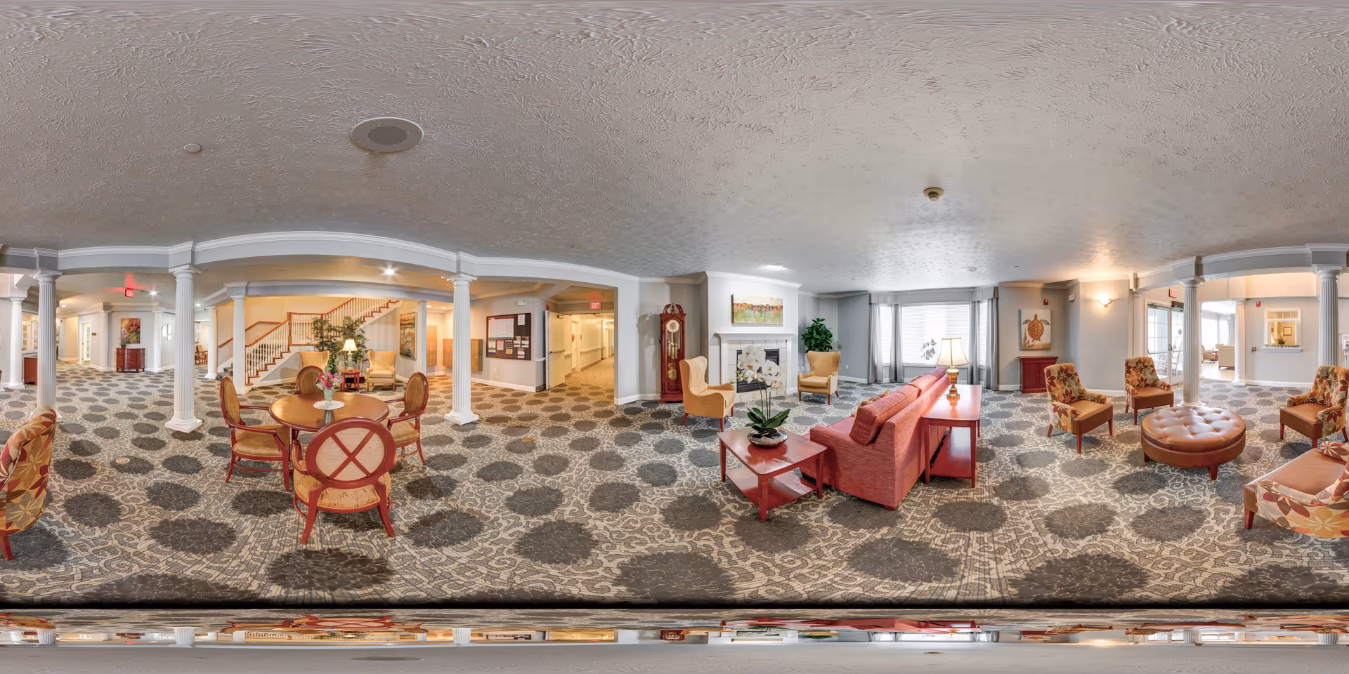 A spacious senior living facility common area with patterned carpet, multiple seating arrangements including armchairs and sofas, a round table with chairs, a grandfather clock, a fireplace with artwork above it, and large windows with curtains allowing natural light.
