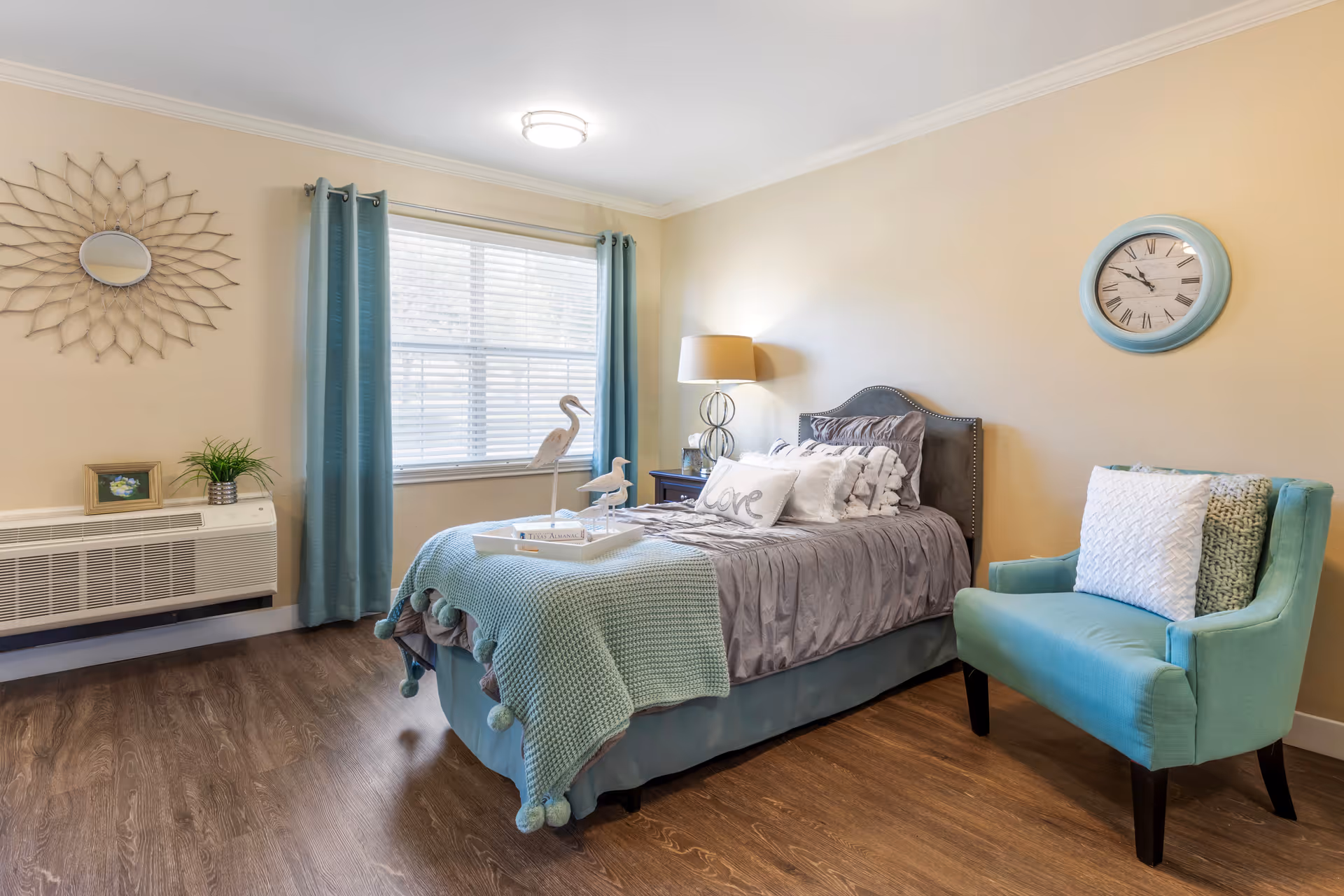 A cozy bedroom with a single bed dressed in gray and teal bedding, including a teal throw blanket with pom-poms and decorative pillows. A tray with two bird figurines and a book sits on the bed. To the right of the bed is a teal upholstered chair with two pillows. The room has wood flooring, beige walls, a window with teal curtains, a round wall clock with a teal frame, a decorative sunburst mirror, a nightstand with a lamp, and a small plant on a heater unit.