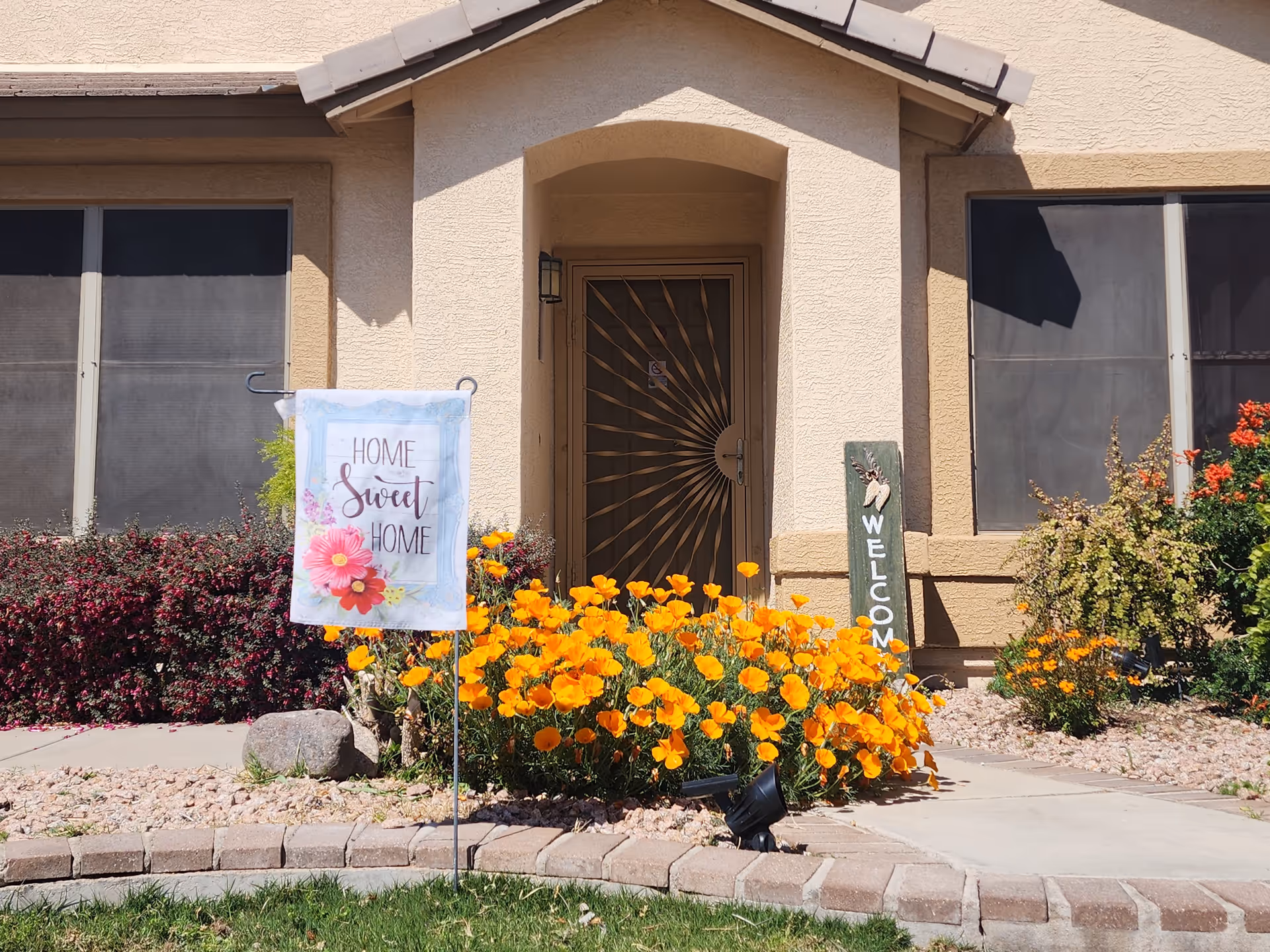 Front entrance of a beige stucco building with a decorative metal security door. There are two large windows on either side of the door with dark screens. In front of the entrance, there is a garden bed with vibrant orange flowers and other greenery. A small sign in the garden reads 'WELCOME' vertically, and another decorative flag sign says 'HOME Sweet HOME' with floral designs. The area is well-lit by sunlight.