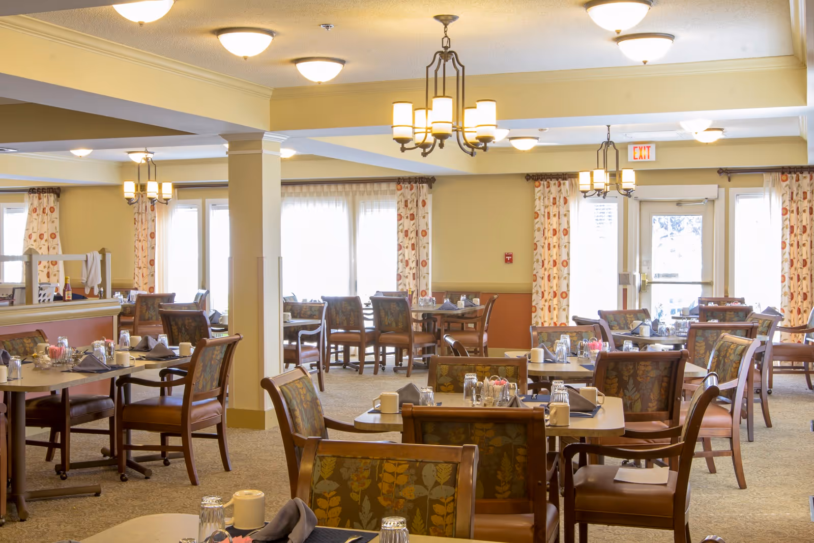 A bright and spacious dining room in a senior living facility with multiple tables and chairs arranged neatly. Each table is set with glasses, napkins, and condiments. The room features large windows with patterned curtains, warm lighting from ceiling fixtures, and a carpeted floor.