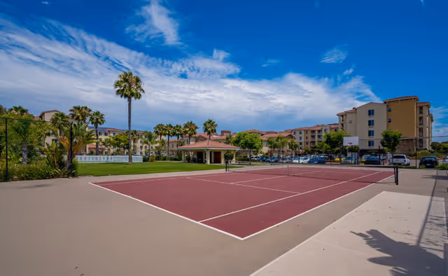 Tennis court on the grounds of a senior living complex with palm trees, a gazebo, and multi-story buildings under a blue sky.