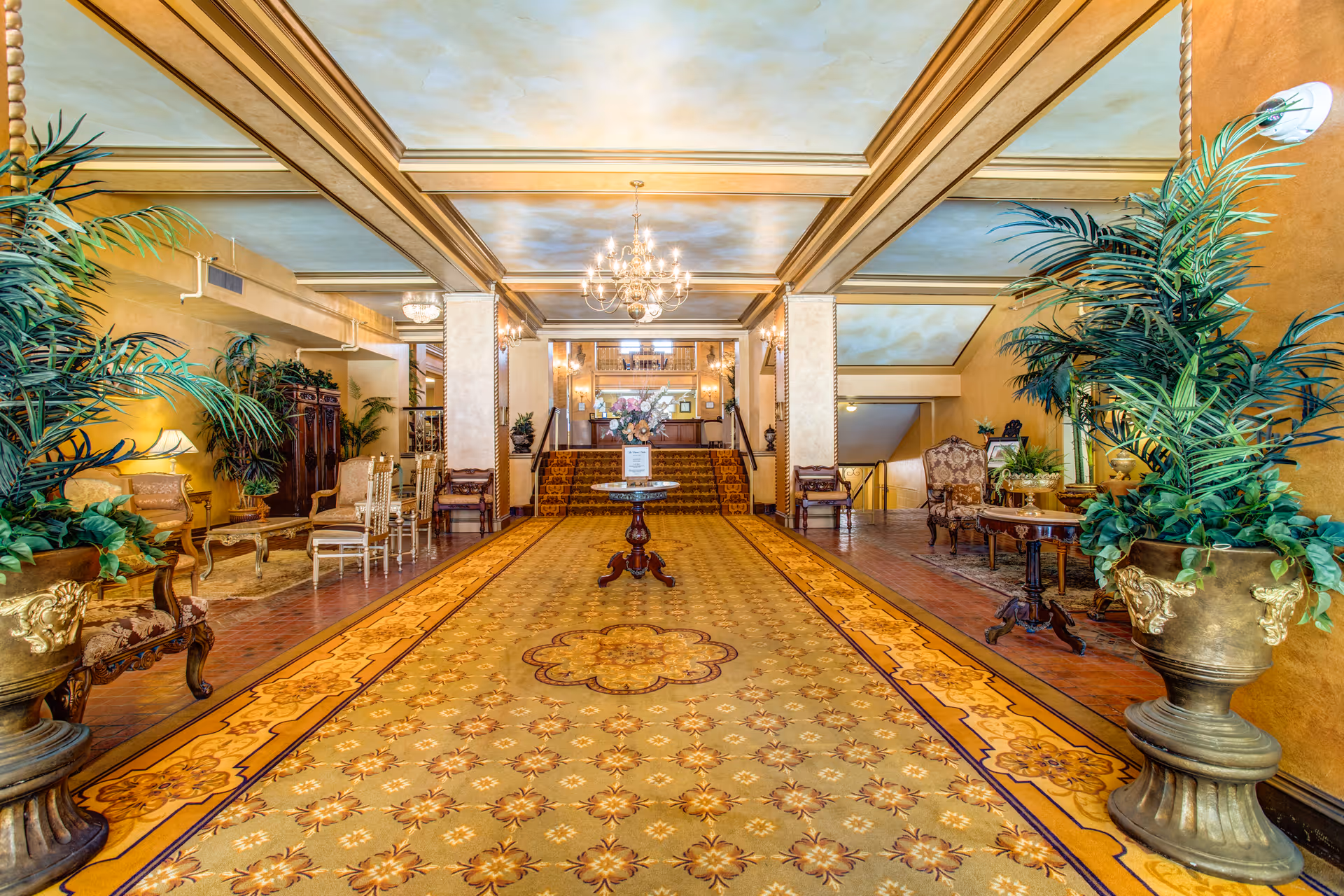 A spacious and elegant lobby area with a patterned carpet, ornate wooden furniture, large potted plants, and a chandelier hanging from a coffered ceiling. There is a round table with a floral arrangement in the center and stairs leading to another level in the background.