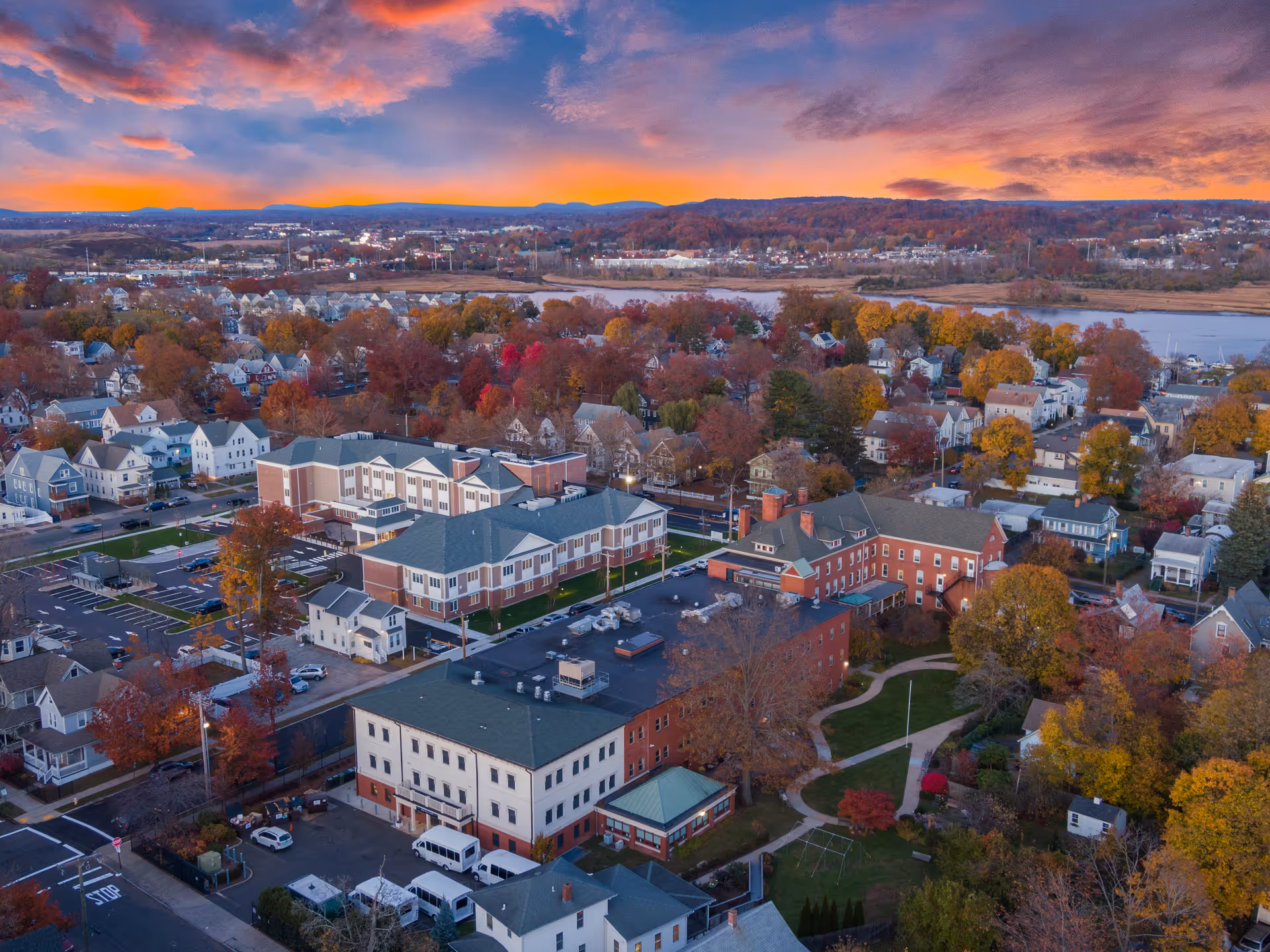 Aerial view of a senior living facility named Mary Wade surrounded by residential houses and trees with autumn foliage under a colorful sunset sky.