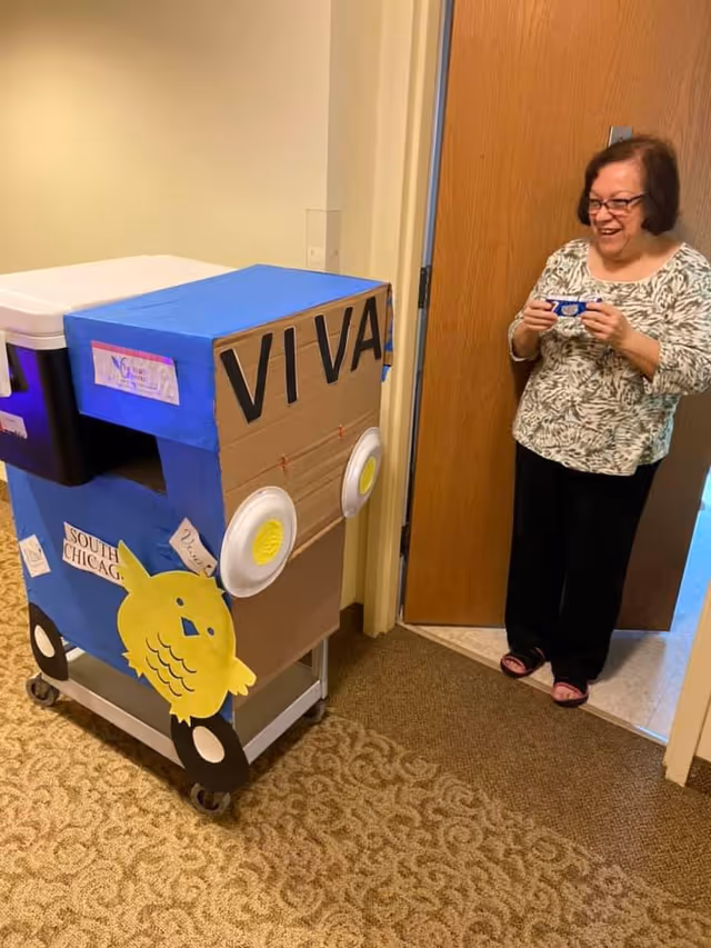 A woman standing in a doorway smiling and holding a small object, next to a decorated cart made to look like a blue vehicle with the word 'VIVA' on the front and a yellow bird cutout on the side. The cart has wheels and is placed on a patterned carpeted floor inside a building.