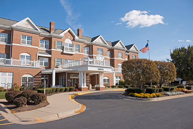Exterior view of a three-story brick senior living facility with white trim and multiple windows. The entrance features a covered drop-off area with columns and the number 16051 displayed. There are landscaped bushes, trees, and flower beds around the driveway, and an American flag is flying on a flagpole near the entrance under a partly cloudy blue sky.