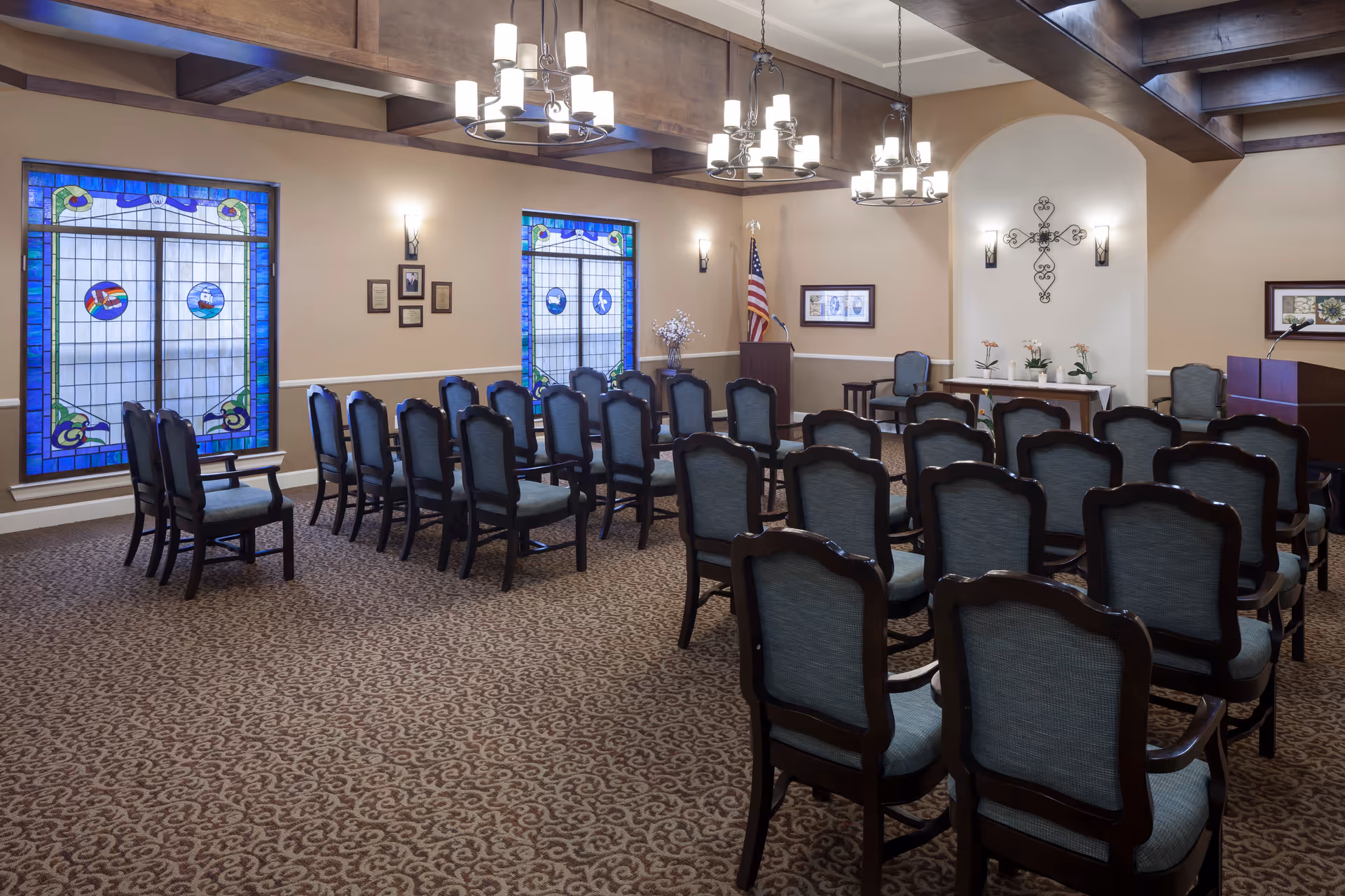 Interior view of a chapel or meeting room with rows of blue upholstered chairs arranged facing a small altar area. The room features stained glass windows with blue borders, beige walls, decorative wall sconces, and three chandeliers hanging from a wooden beam ceiling. There is an American flag near a podium and a decorative cross on the wall behind the altar.