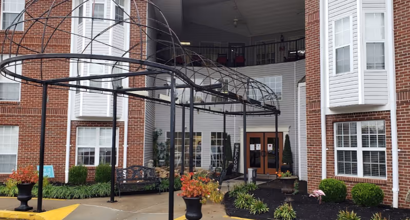 Covered metal pergola and landscaped entrance to a multi-story brick senior living building with double glass doors.