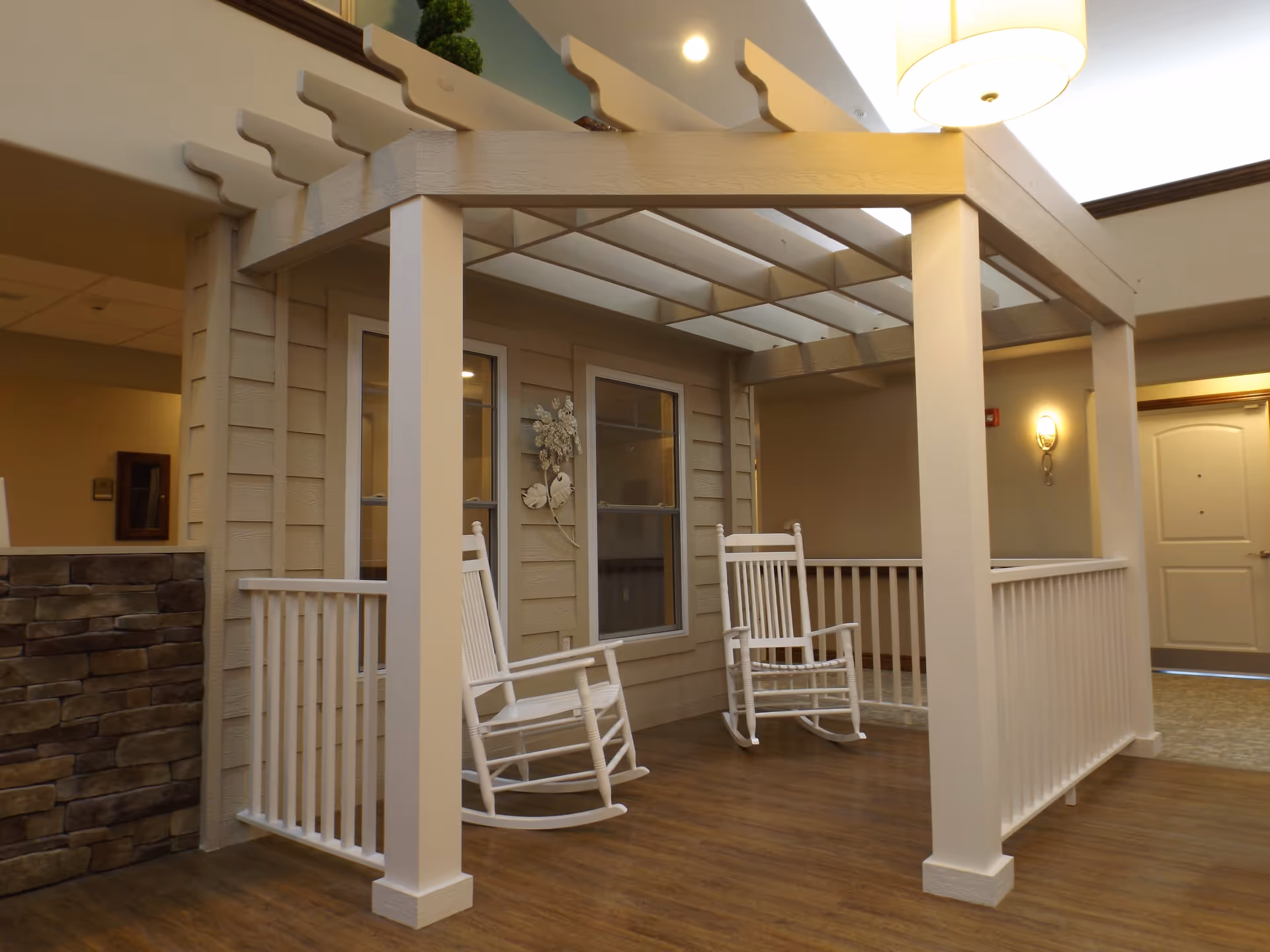 Indoor seating area with two white rocking chairs under a white pergola structure, beige walls with two windows, a decorative wall piece, and wood-style flooring in a senior living facility.