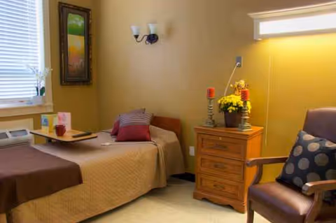 Cozy bedroom in a senior living facility featuring bed, dresser, and chair.