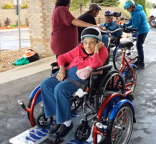 An older woman wearing a helmet and pink jacket sits in a wheelchair attached to an adaptive tricycle while others adjust bicycles under a covered entrance.