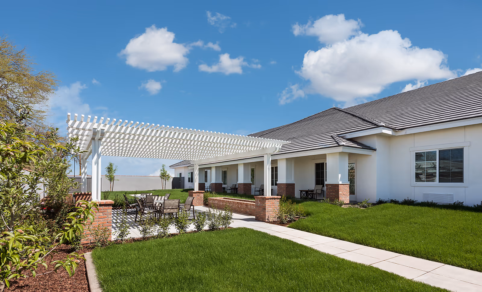 Sunlit outdoor courtyard of a white assisted living building with a pergola, patio seating, and green lawn.