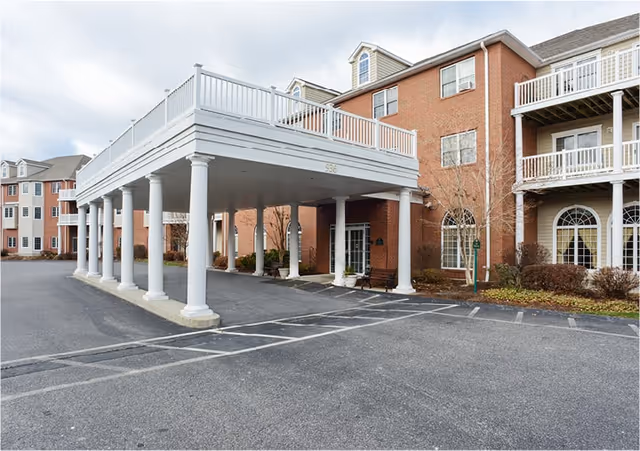 Exterior view of a senior living facility with a large covered entrance supported by white columns. The building is made of red brick with white balconies and windows. There is a parking area in front of the entrance and some landscaping with bushes and small trees around the building.