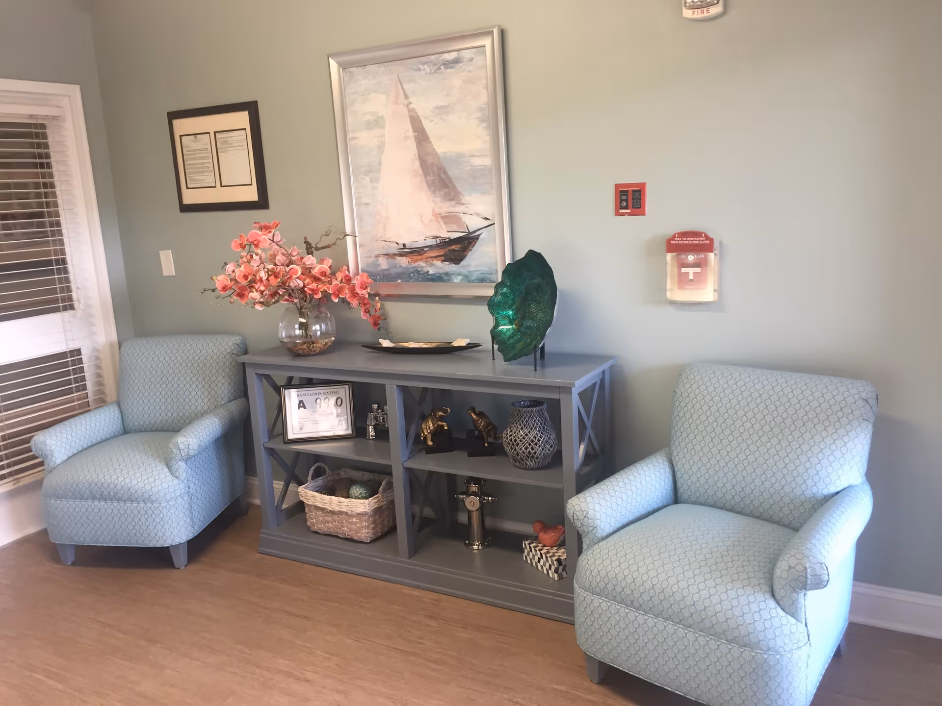 A cozy seating area with two light blue upholstered armchairs on either side of a gray console table. The table holds decorative items including a vase with pink flowers, a framed certificate, small sculptures, and a basket. Above the table is a framed painting of a sailboat on the water. The walls are painted light green and there is a fire alarm and fire safety equipment mounted on the wall.
