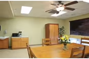A dining area in a senior living community featuring a wooden table with chairs, a vase of yellow flowers on the table, a ceiling fan with lights, a wooden cabinet, a small sink, a microwave, and a coffee maker against a light green wall with a window covered by dark blinds.