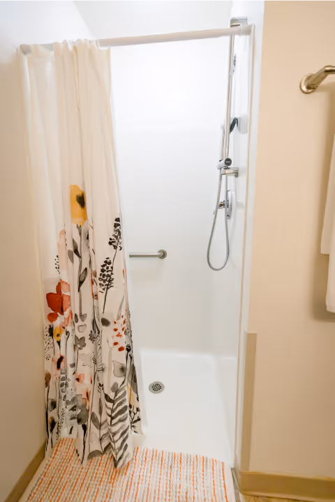 Small white shower stall with a floral shower curtain, handheld showerhead, grab bar, and striped bath mat.