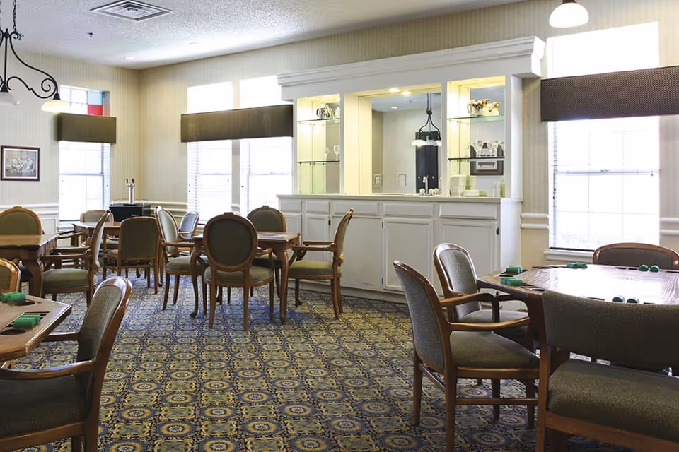 A bright dining/activity room with multiple wooden tables and upholstered chairs and a built-in serving cabinet.