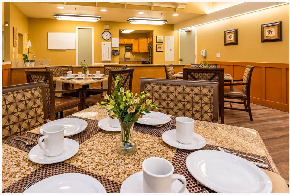 A dining area in a senior living facility with tables set with white plates, cups, and silverware. The tables have patterned placemats and a small vase with flowers as a centerpiece. The room has warm yellow walls, wooden paneling, and framed artwork. In the background, there is a kitchen area with wooden cabinets and a clock on the wall.