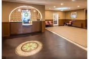 Reception area of Highland Rehabilitation & Nursing Center featuring a wooden front desk with a decorative arch and a floral design behind it. The floor has a circular floral pattern near the entrance, and there are chairs and a small table along the beige walls in the background.