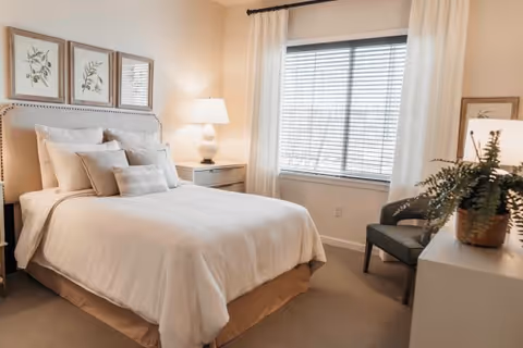A cozy bedroom in a senior living facility featuring a neatly made bed with white linens and multiple pillows. There is a bedside table with a lamp, a window with blinds and white curtains, a comfortable chair, and a dresser with a potted plant. The walls are decorated with framed botanical prints.