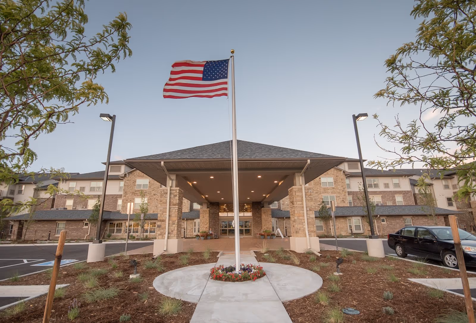 Front exterior view of Parkside Village Retirement Resort featuring a large covered entrance with stone pillars, an American flag on a flagpole in the center, landscaped garden beds, and a parking area with cars on either side.