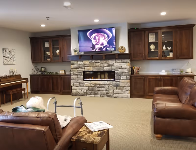A cozy living room with brown leather sofas, a stone fireplace with a modern gas insert, and a flat-screen TV mounted above it showing a black and white image of a man in a hat. The room has built-in wooden cabinets on either side of the fireplace, a piano with a bench on the left, and a small side table with newspapers. An elderly person with white hair is seated on one of the sofas using a walker.