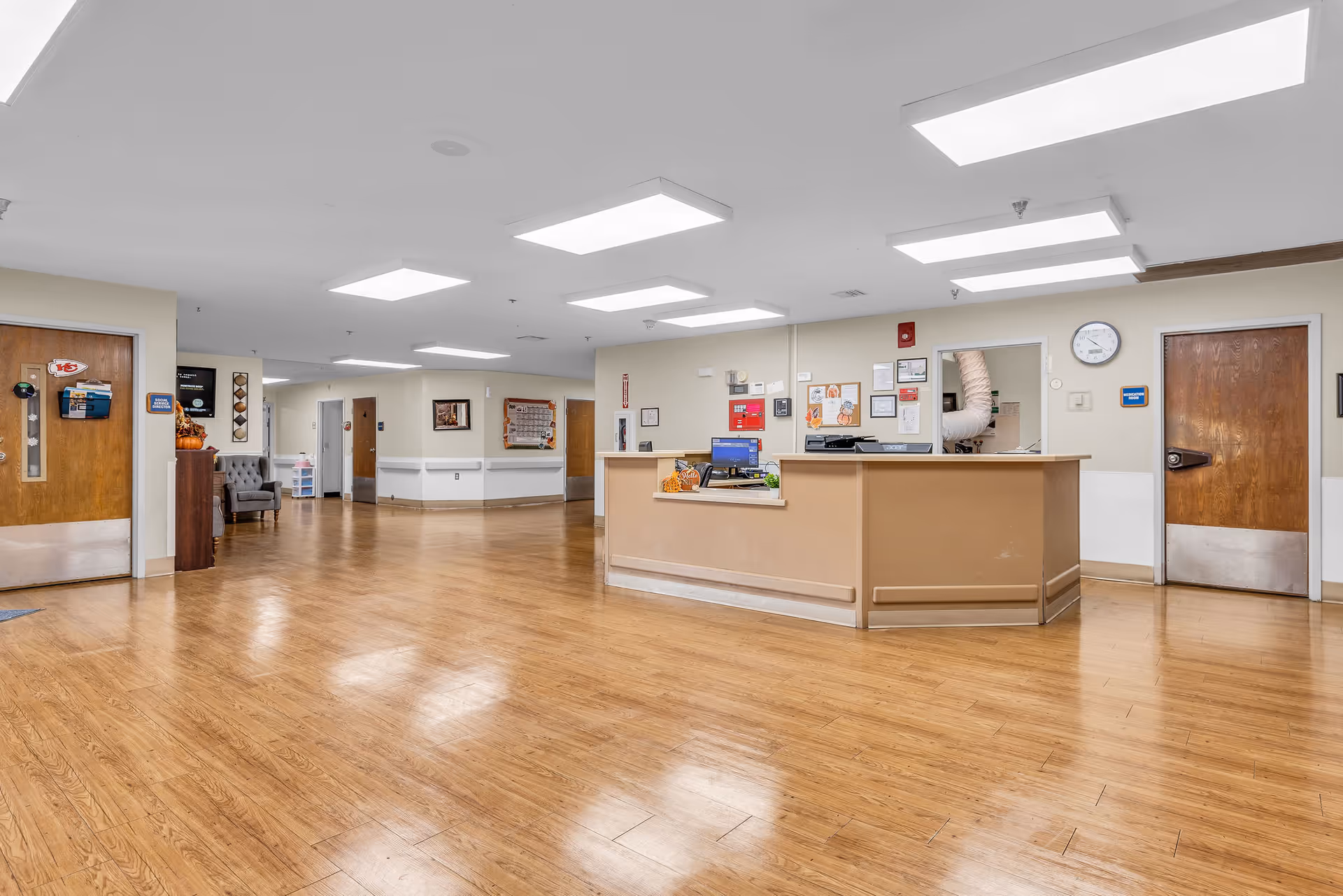 A spacious reception area in a healthcare and rehabilitation facility with a wooden floor, a beige reception desk with computer monitors, and several wooden doors along the walls. The ceiling has multiple rectangular fluorescent lights, and there are chairs and framed pictures on the walls.