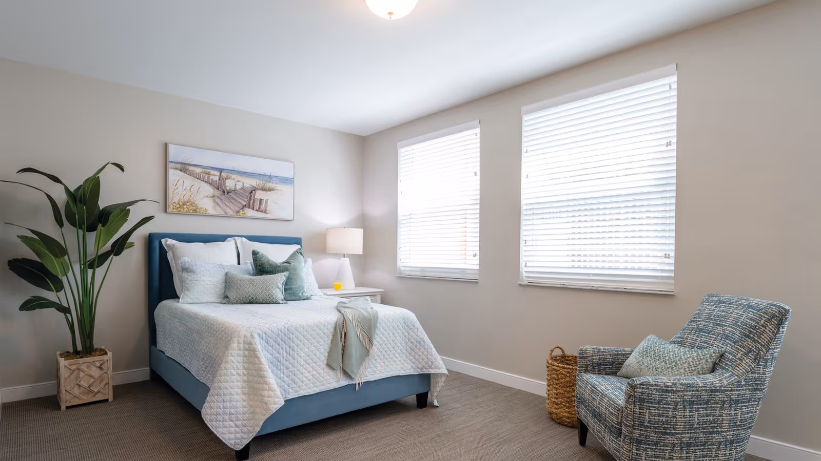Bright bedroom with a blue upholstered bed, white quilt, decorative pillows, a potted plant, wicker basket, and a blue chair by two windows.