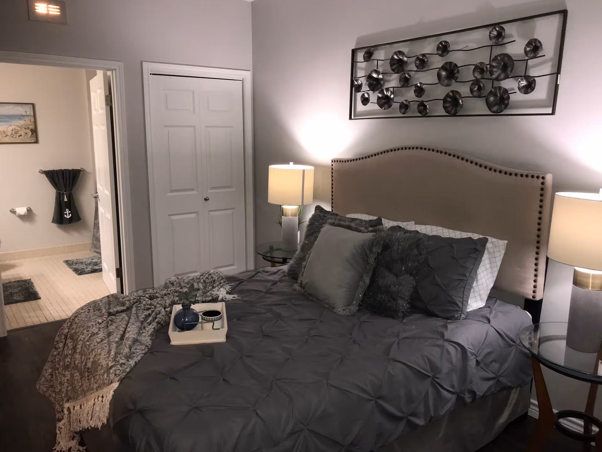 A cozy bedroom with a neatly made bed featuring gray bedding and multiple pillows. Two bedside tables with lamps flank the bed. Above the headboard is a decorative metal wall art piece. In the background, an open door reveals a bathroom with a towel hanging on the wall and a framed picture.