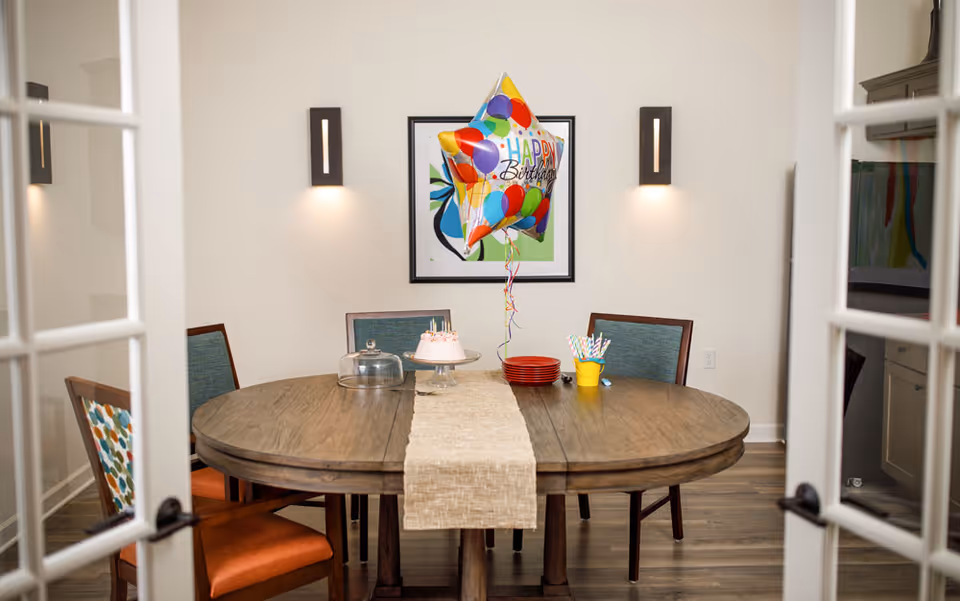 A dining room with a round wooden table set for a birthday celebration. On the table, there is a small birthday cake on a stand, a colorful 'Happy Birthday' balloon, a stack of red plates, and a yellow container holding striped straws. Four chairs with different upholstery surround the table, and the room features wooden flooring, beige walls, two wall sconces, and a framed abstract artwork.