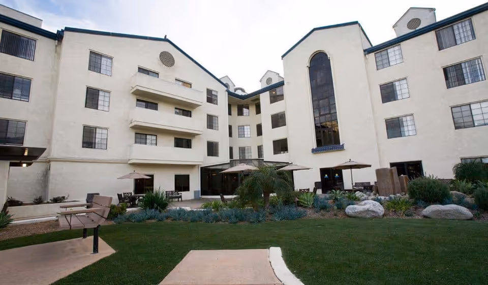 Outdoor courtyard area of a senior living facility with green lawn, benches, umbrellas, and plants in front of a multi-story beige building with multiple windows and balconies.