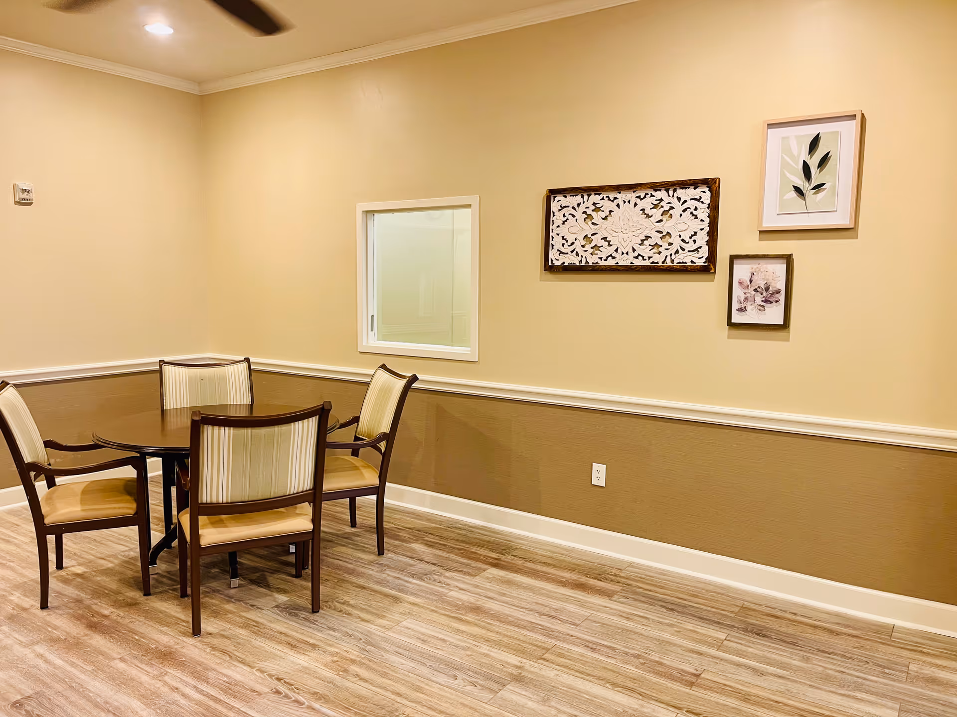 A small dining area with a round wooden table and four cushioned chairs with wooden frames. The walls are painted beige with a white chair rail and three framed decorative artworks hanging on the wall. The floor is light wood, and a ceiling fan is partially visible at the top left corner.