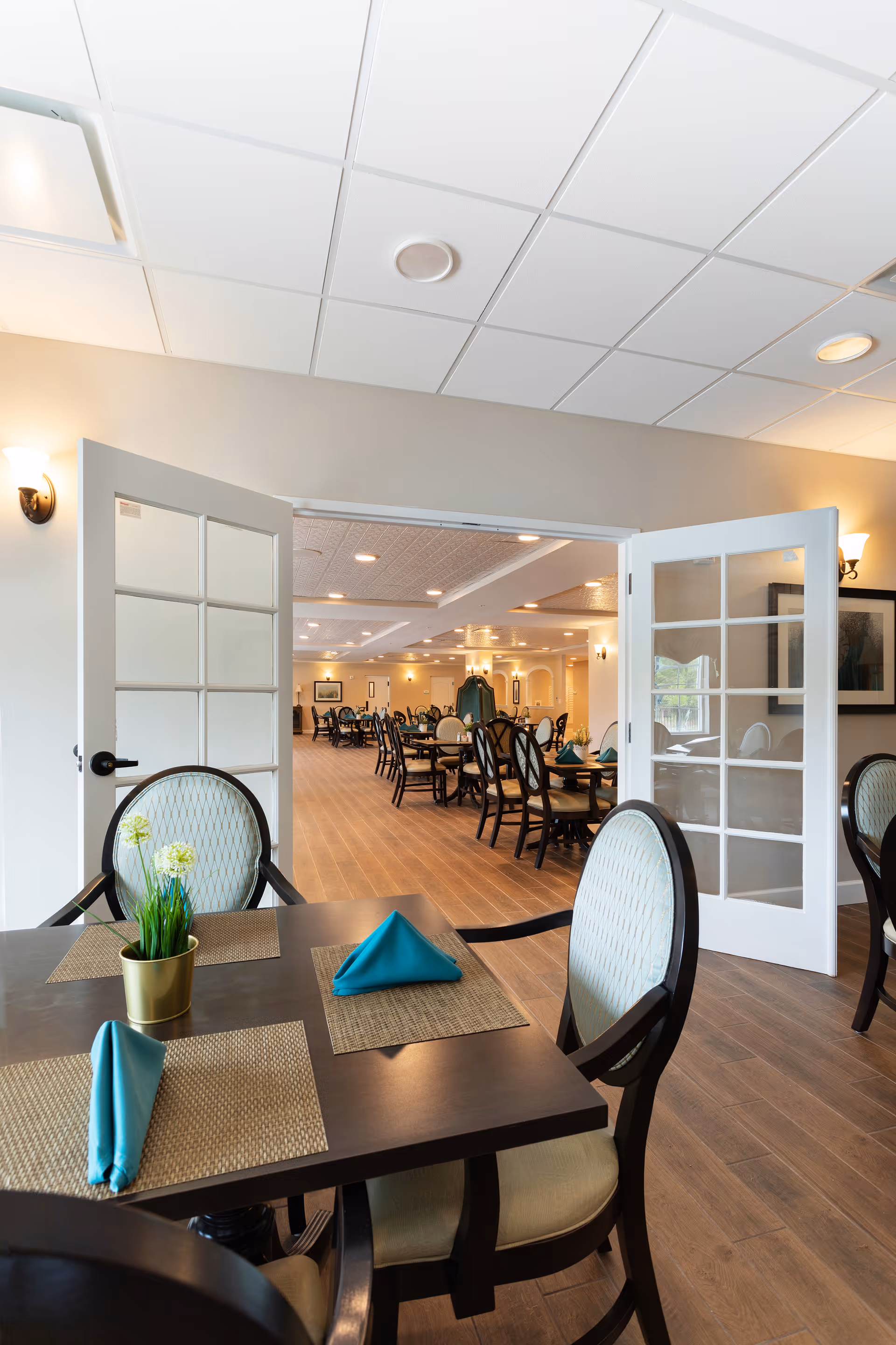 View of a dining area in an assisted living facility with a dark wooden table set for two, featuring teal folded napkins on woven placemats and a small potted plant. The room has light-colored walls, wood flooring, and white French doors opening into a larger dining room with multiple tables and chairs arranged neatly under recessed lighting.