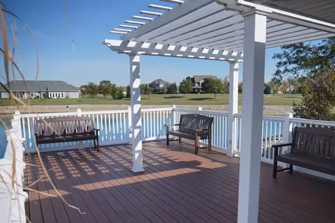 Wooden deck with a white pergola and benches overlooking a pond and distant houses under a clear sky.