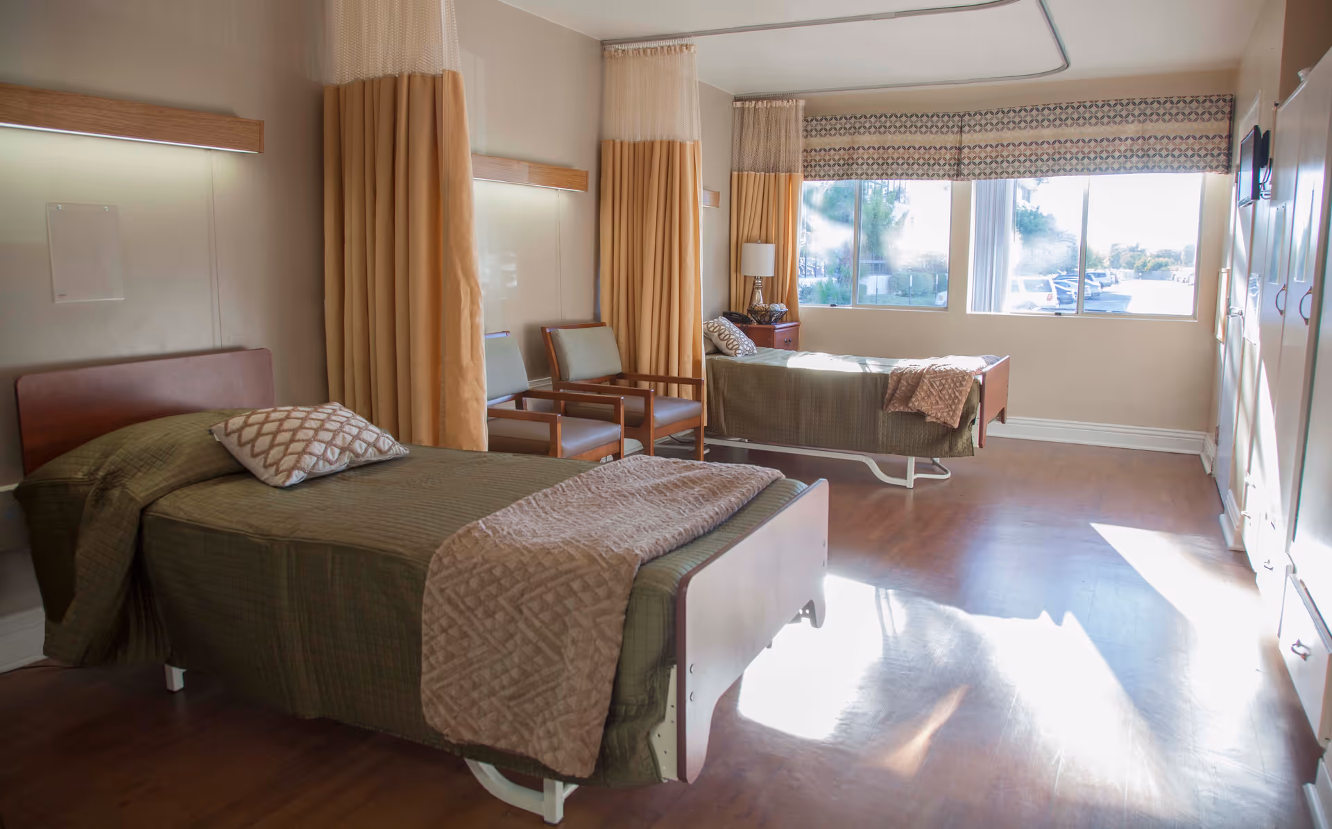 A bright and clean room in a healthcare facility with two hospital-style beds, each with green bedding and a patterned pillow. There are two chairs between the beds, beige curtains for privacy, a large window letting in natural light, and a small bedside table with a lamp. The floor is wooden, and there are cabinets along one wall.