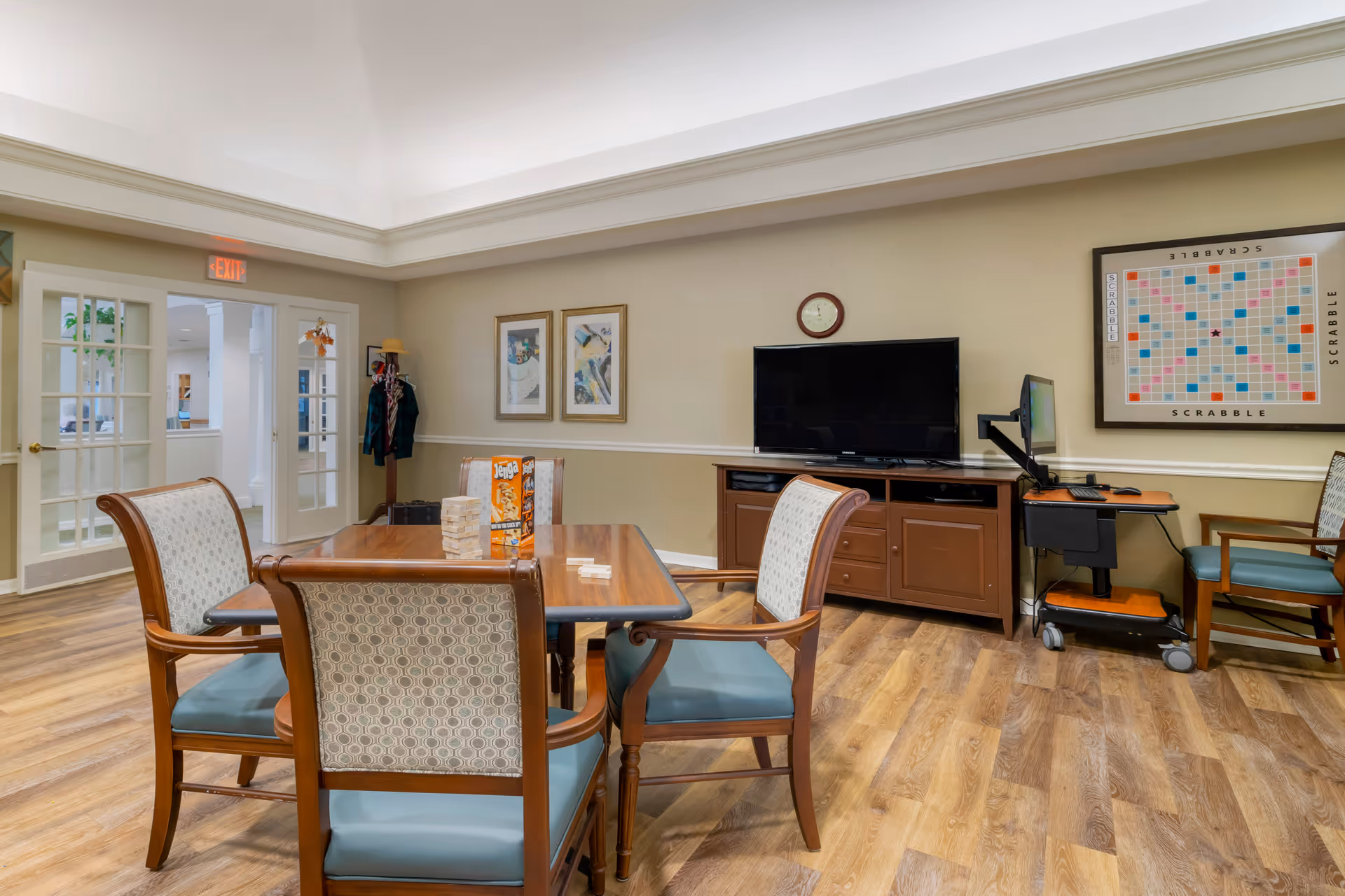 Interior common room with a wooden table and chairs, a television on a cabinet, and a Scrabble board on the wall.