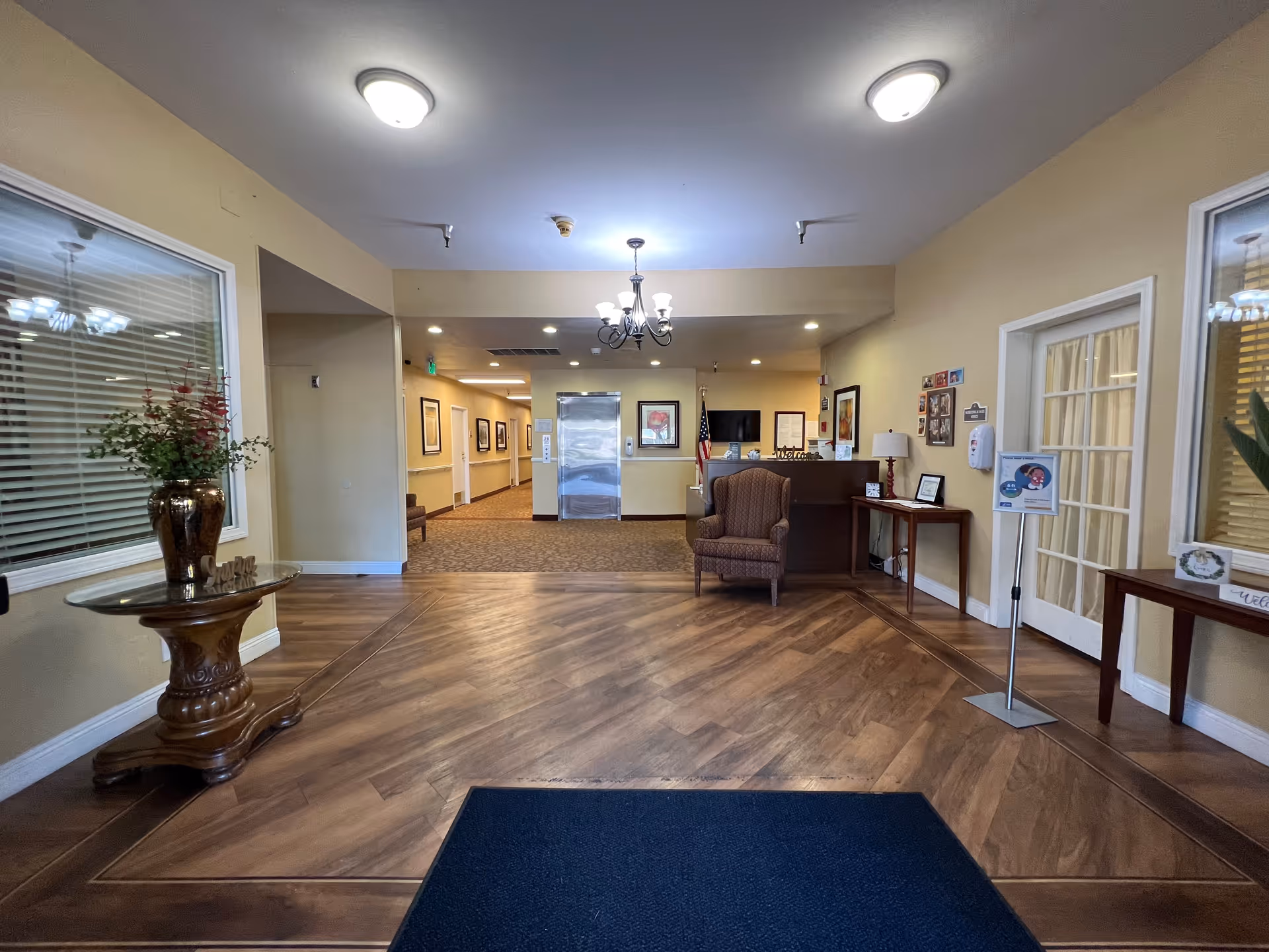 Interior view of a senior living facility lobby with wooden flooring, a small reception desk, an armchair, decorative tables with plants, framed pictures on the walls, and ceiling lights. There is a hallway leading further into the building and an elevator at the end of the hallway.