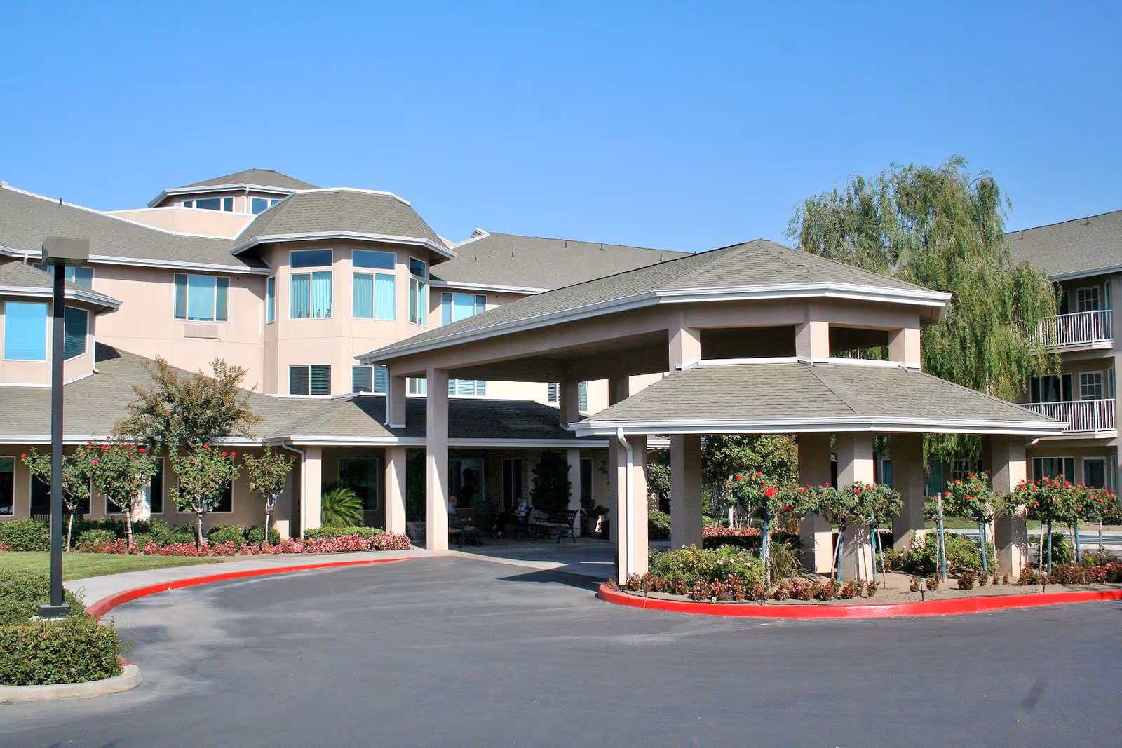 Front entrance of a multi-story senior living building with a covered porte-cochere, landscaped beds, and a clear blue sky.