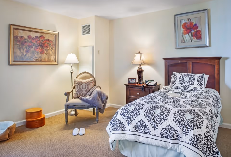 A cozy bedroom with a single bed covered in a white and black patterned comforter. Next to the bed is a wooden nightstand with a lamp, a framed photo, and a telephone. A cushioned chair with a pillow and a blanket is placed near a floor lamp. The walls are decorated with two framed floral paintings. A pair of white slippers is on the carpeted floor near the chair.
