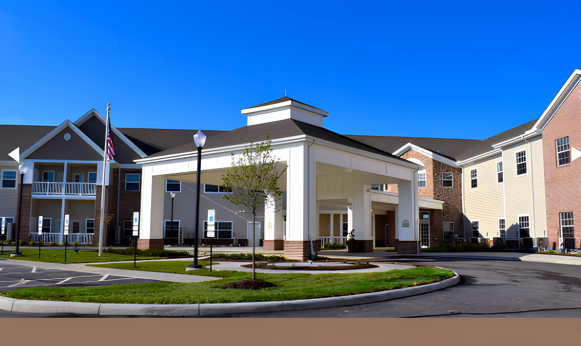 Exterior view of Danbury Huber Heights senior living facility showing the main entrance with a covered drop-off area, landscaped grounds, an American flag, and a clear blue sky.