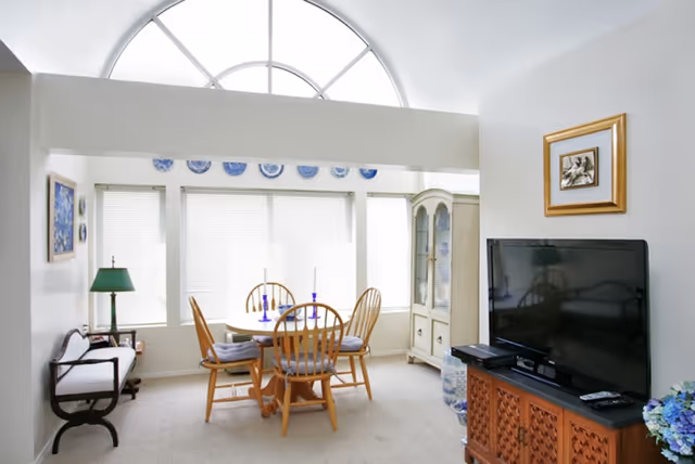 A bright room with a round wooden dining table surrounded by four wooden chairs with cushions. Behind the table are three large windows with blinds and a decorative row of blue and white plates above them. To the right is a white display cabinet with glass doors and blue decorative items inside. On the right side of the image is a flat-screen TV on a wooden cabinet with a framed picture hanging above it. On the left side, there is a wooden bench with a white cushion and a green table lamp on a small side table next to it. The room has a high ceiling with a large arched window letting in natural light.