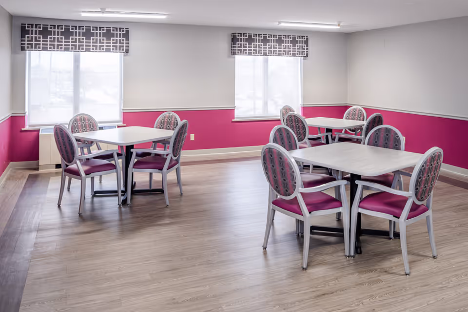 Bright dining room with several square tables and pink-accented chairs against windows and pink lower walls.
