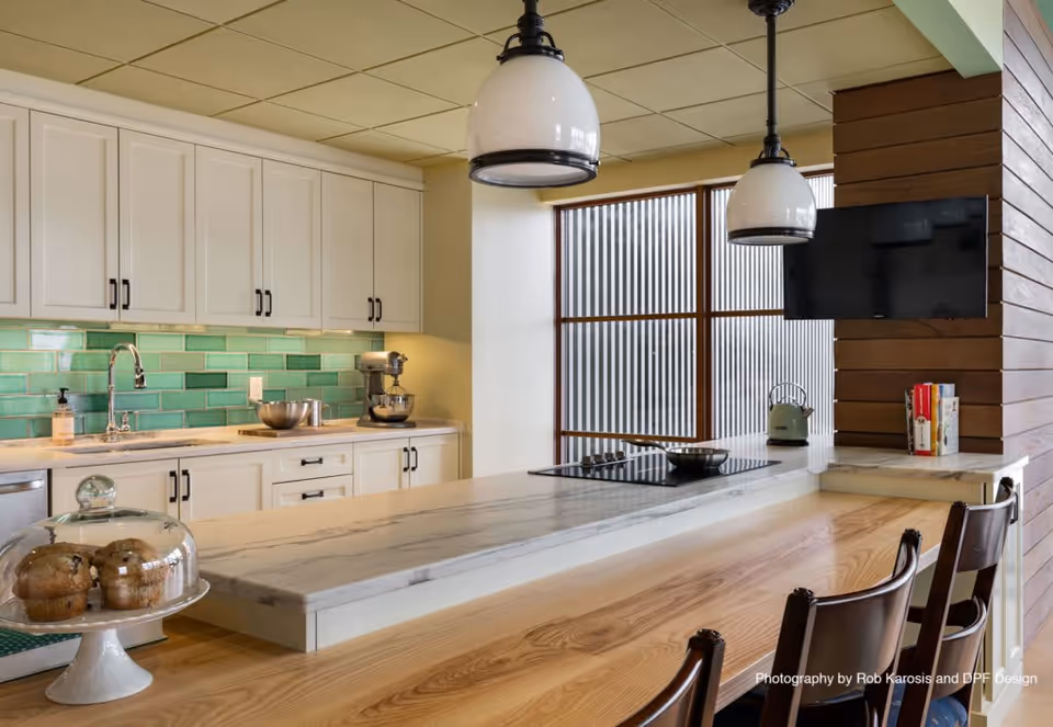 Modern kitchen area with white cabinets, a green tiled backsplash, a marble countertop island with a built-in stove, two hanging pendant lights, a glass dome with muffins on a stand, and a small television mounted on a wooden accent wall.