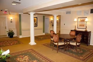 Interior view of a common area in a senior living facility with beige walls, carpeted floors, decorative rugs, white columns, a round wooden table with four upholstered chairs, framed artwork on the walls, and a sideboard with two lamps.